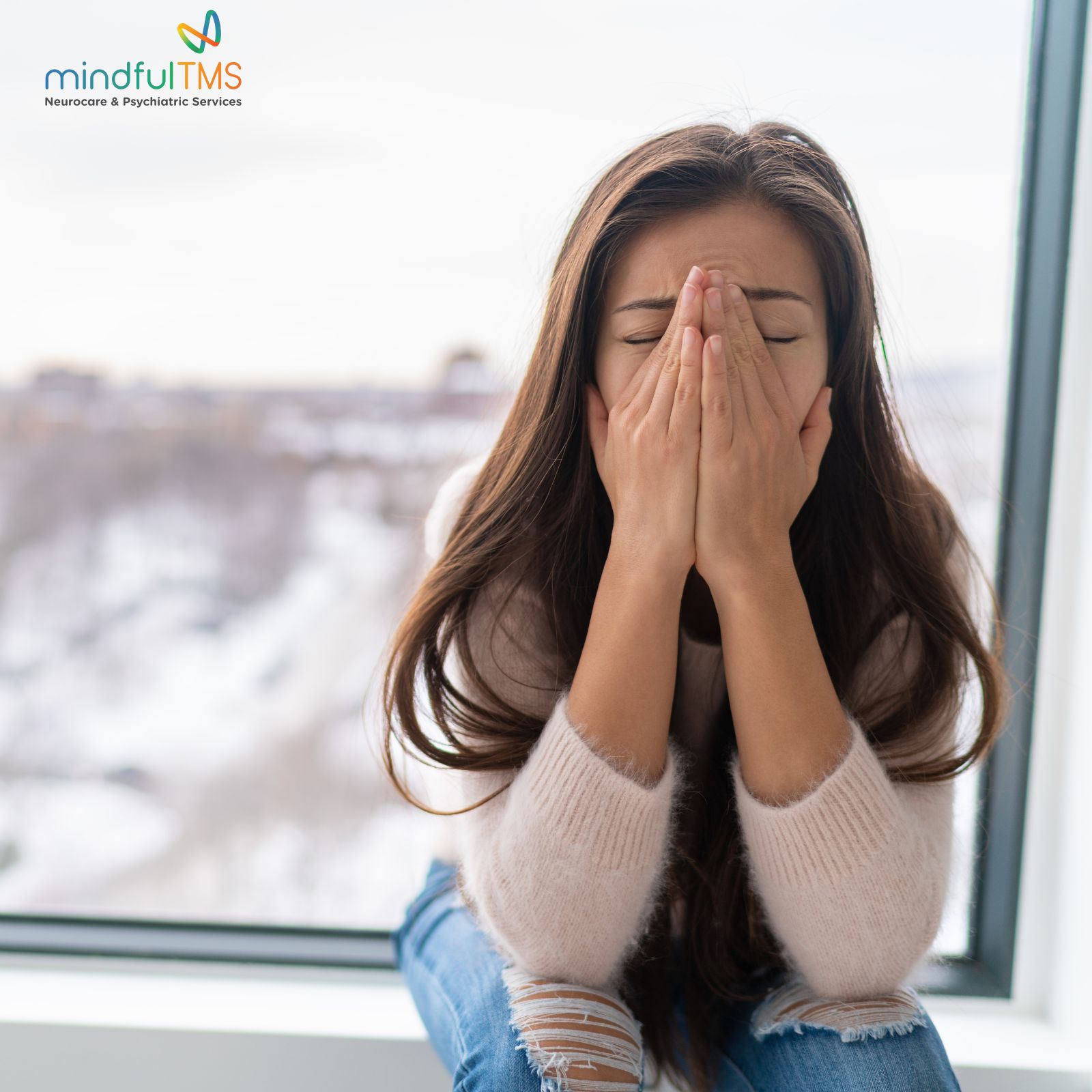 Woman sitting by a window with her hands to her face, snowy landscape in the background.