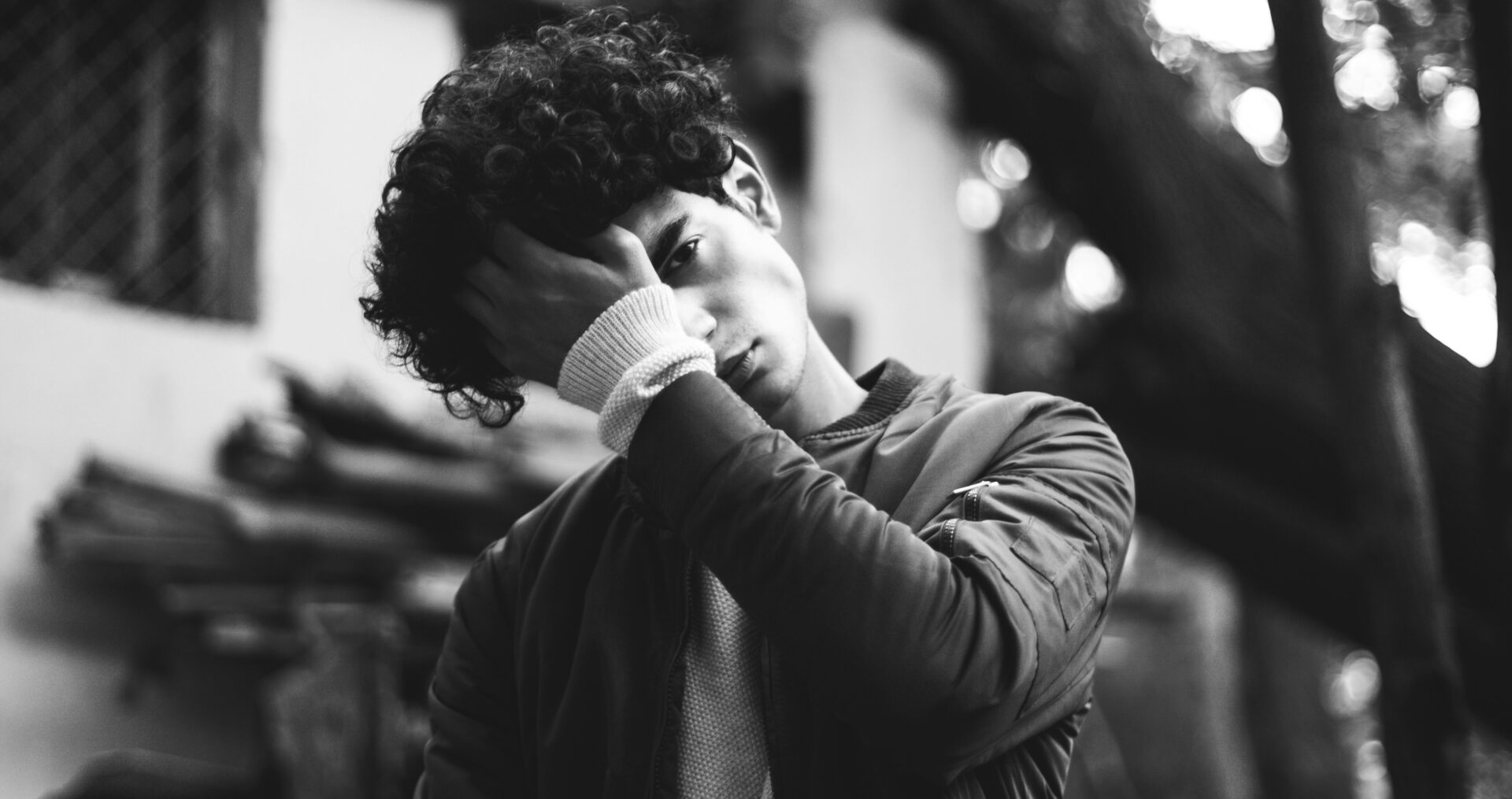A young man with curly hair in a pensive, emotional state, holding his head with one hand, representing the deep emotional and physical pain of heartbreak. The black-and-white aesthetic enhances the raw, introspective mood.