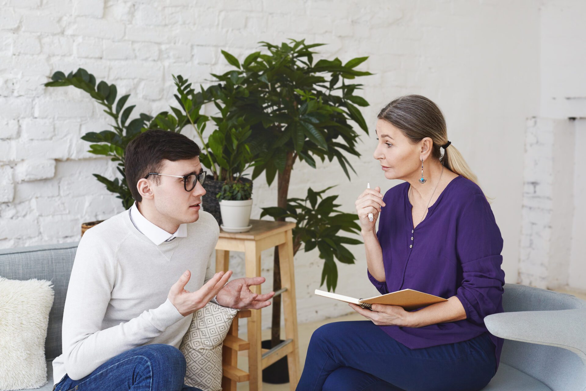 A young man wearing glasses and a light-colored sweater sits on a couch, gesturing with his hands as he speaks to a middle-aged female therapist in a purple blouse. She holds a notebook and pen, listening attentively. The setting is a cozy, modern therapy office with a white brick wall and green plants in the background.