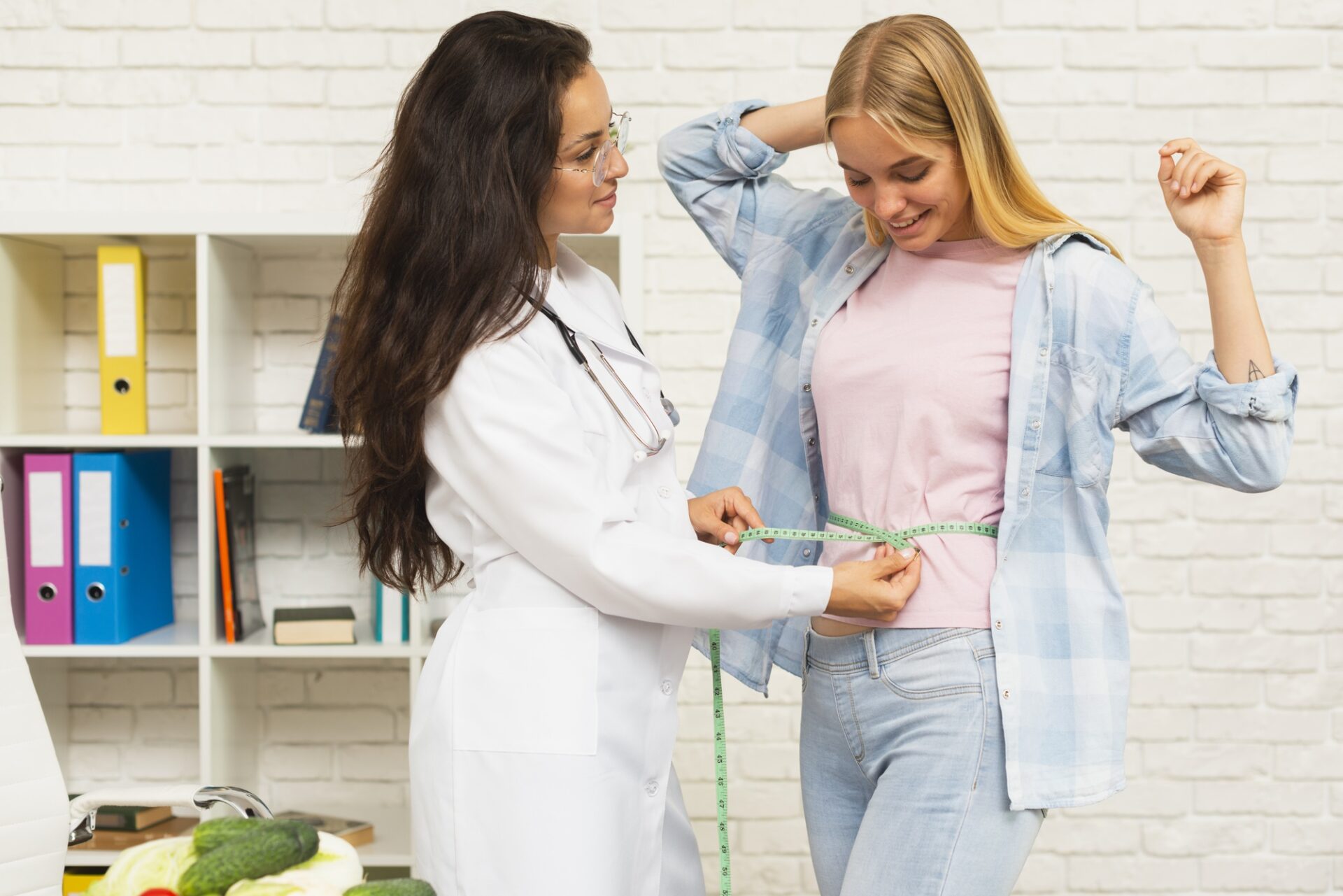 A smiling woman in a pink shirt and blue flannel being measured around her waist by a doctor, symbolizing potential benefits of TMS therapy for weight loss.